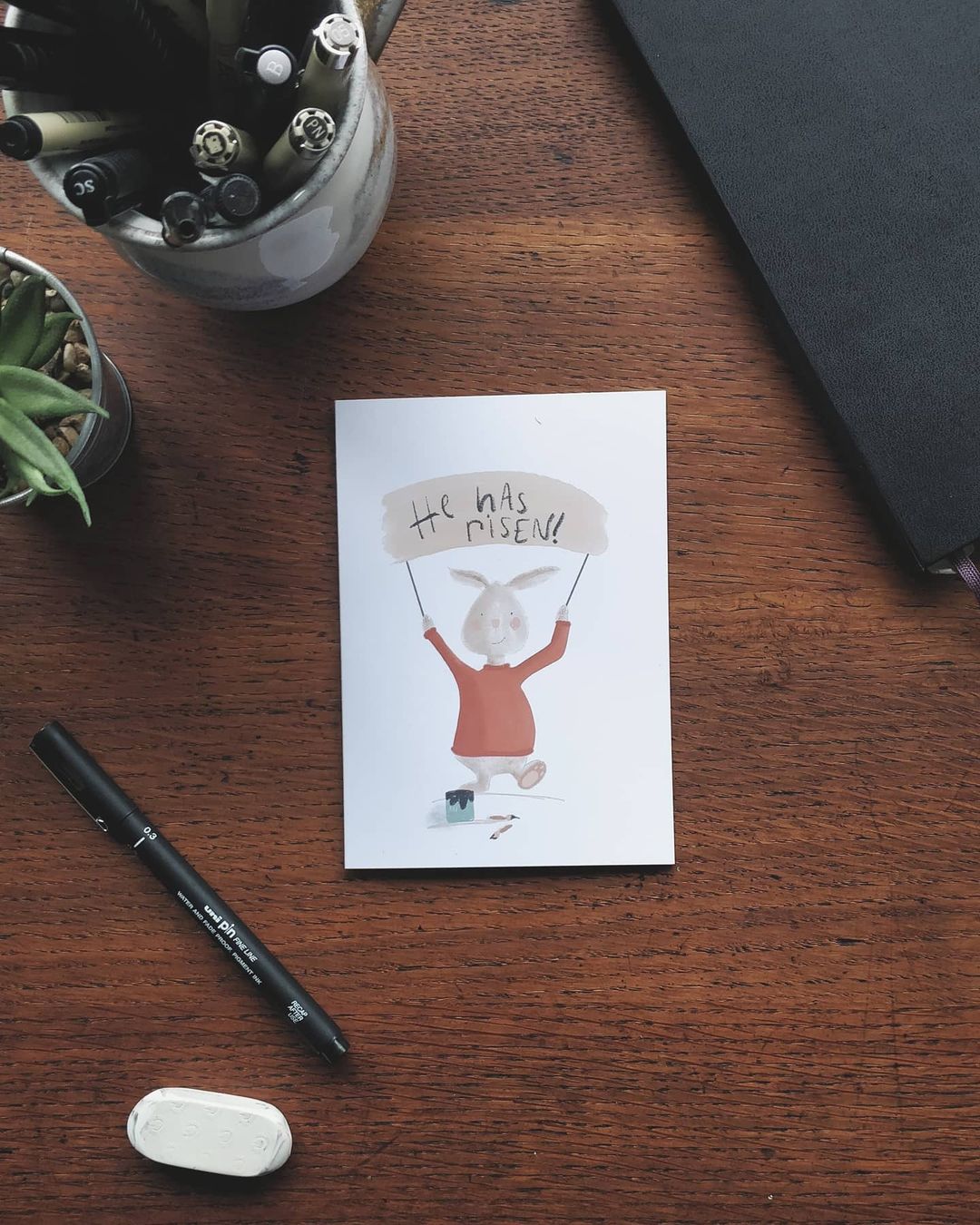 An image of an Easter Card with a rabbit holding a sign saying 'He Has Risen', illustrated by Hello You Create.